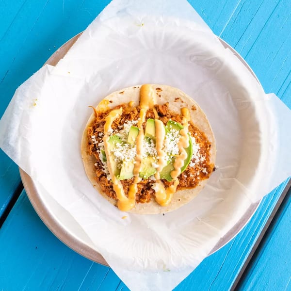 A single taco with shredded meat, avocado slices, crumbled cheese, and orange sauce on a blue table.