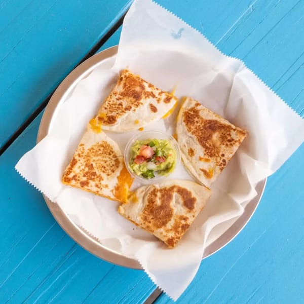 Four quesadilla slices with melted cheese and a side of guacamole on a metal plate.
