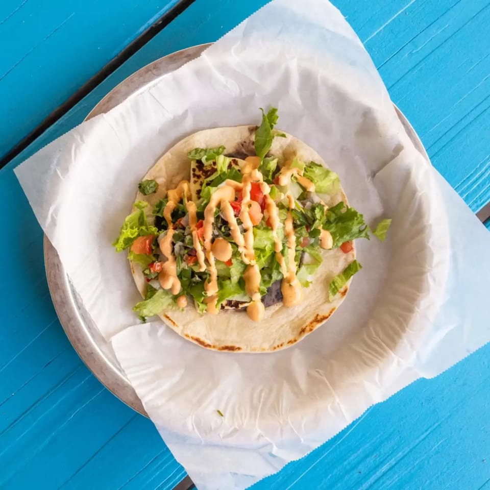 A single taco with lettuce, tomatoes, and sauce on a metal plate with white paper, on a blue table.