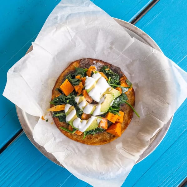 A tostada with spinach, sweet potato, avocado, and a white sauce on a metal plate.